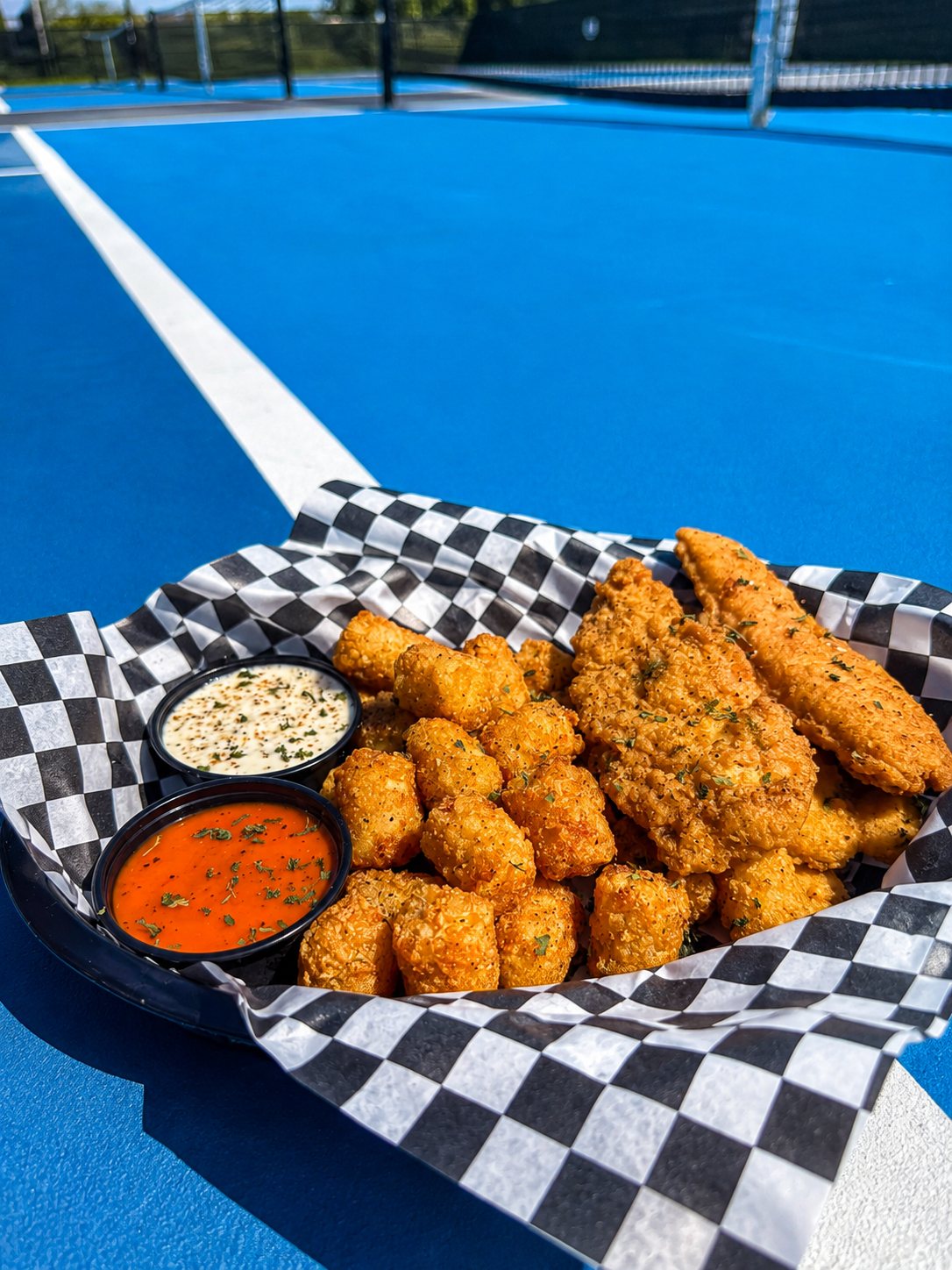 A basket of crispy chicken tendies and Yacht Tots with two dipping sauces, courtside on the pickleball court